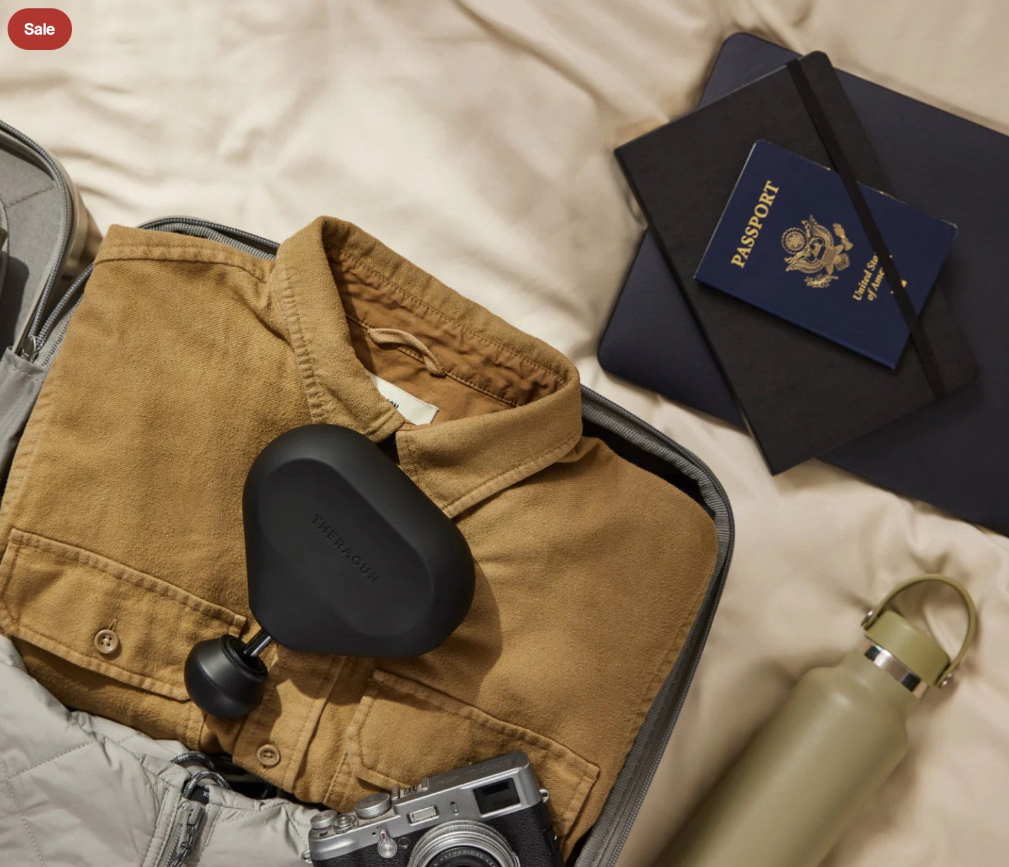 Theragun Mini resting on a folded shirt in a suitcase alongside a passport, water bottle, and camera