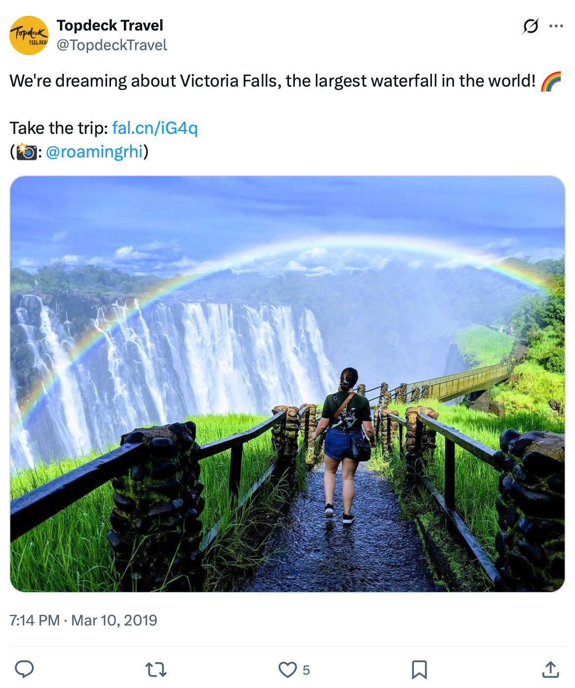 Person walks along a misty wooden trail toward Victoria Falls with a vibrant rainbow arching across the sky
